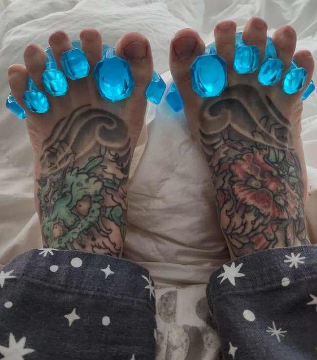 Feet with tattoos and blue pedicure separators, wearing star-patterned pants on a bed
