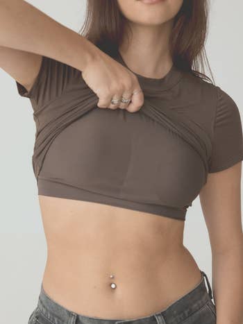 Person lifting a fitted, short-sleeve apical  to uncover  a belly fastener  ringing  and jeans, showcasing the garment's flexibility and fit