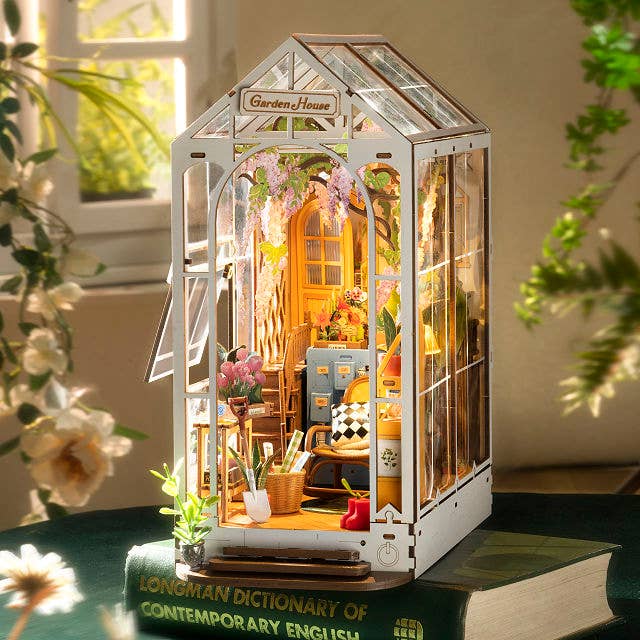 Miniature greenhouse model with intricate interior details placed atop a book, surrounded by greenery, creating a cozy, inviting scene