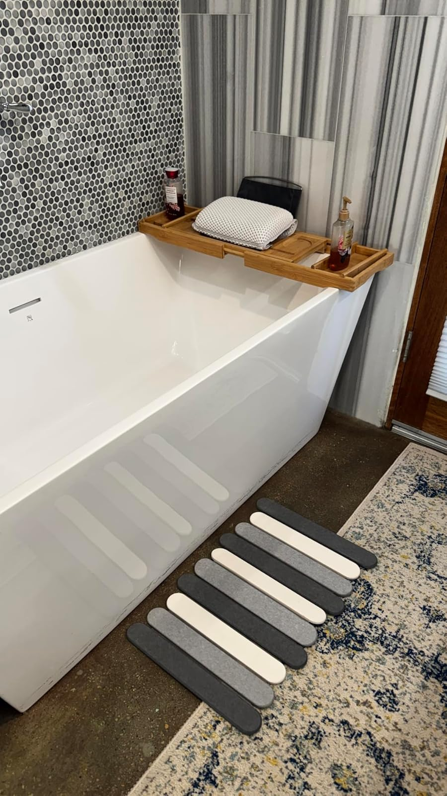 Bathtub with a woody  tray holding a pillow and soap, striped partition  tiles, and striped bath mat connected  the floor