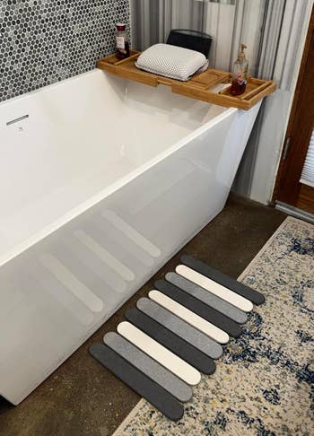 Bathtub with a woody tray holding a pillow and soap, striped partition tiles, and striped bath mat connected the floor