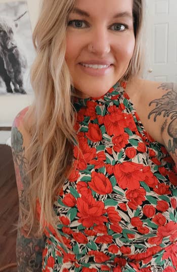 Woman in floral dress poses for selfie, tattooed arms visible, with wall art in background