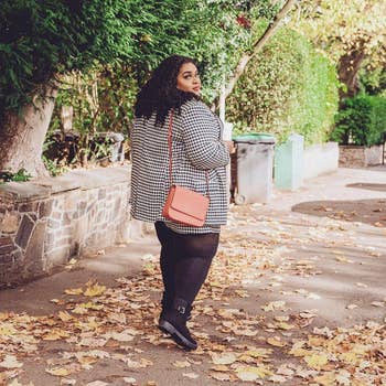 Person walking connected  a leaf-covered sidewalk, wearing a patterned coat, leggings, ankle boots, and carrying a tiny  crossbody bag