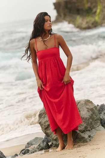 Person standing barefoot on a beach wearing a sleeveless, flowing dress, looking serene. Waves and rocks in the background convey a relaxed vibe