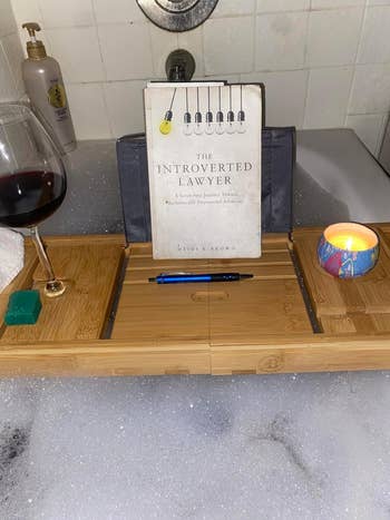 A book titled 'The Introverted Lawyer' on a bathtub caddy with a glass of wine, candle, and toiletries in a foamy bath