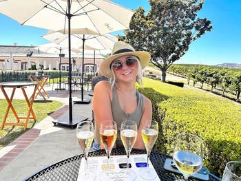 Woman in sun hat sitting at table with glasses of wine, outdoors at a vineyard