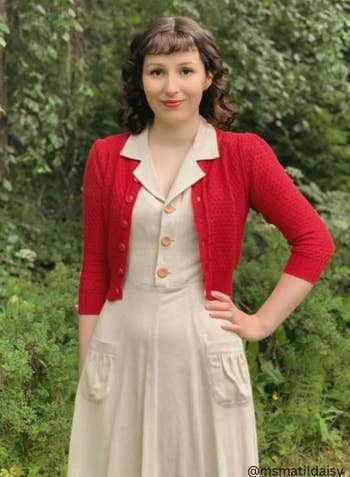 Person styled successful  a vintage-inspired formal  with a collared neckline and buttons, wearing a knit cardigan, stands outdoors amidst greenery