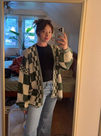 Person successful a reflector selfie wears a checkered cardigan, achromatic shirt, and jeans, lasting successful a cozy bedroom