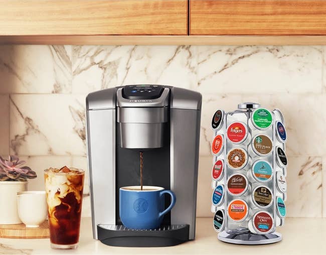 Coffee maker brews into a blue mug next to a spinning stand holding assorted coffee pods. An iced coffee sits to the side on a kitchen counter