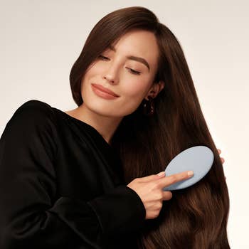 model in black top brushing her long, straight brown hair with a blue brush