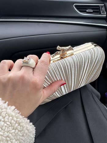 A hand with red nails holds a pleated, metallic clutch in a car, showcasing an engagement ring with a large stone