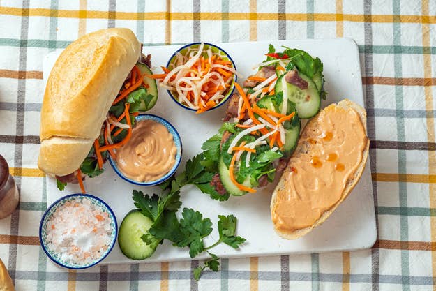 Bánh mì-Sandwiches mit Gurke, Karotten, Koriander und Soßen auf einem weißen Brett über einer karierten Tischdecke