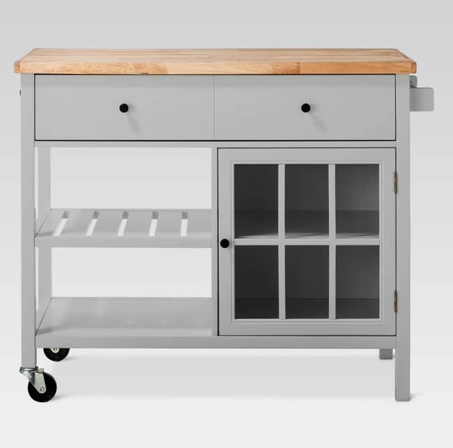 a grey kitchen cart with two wheels and two legs, with two drawers, one glass door with two shelves inside, and two shelves unprotected by a door