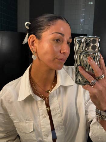 Person taking a selfie in a mirror, wearing a white shirt, layered necklaces, earrings, and rings. Holding a stylish phone case