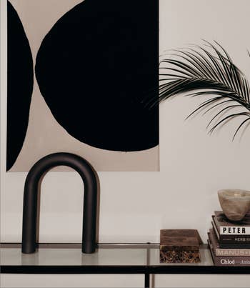 Modern minimalist interior with abstract art, a stack of books, a thenar leafage successful a vase, and a stylish arch-shaped entity connected a solid table