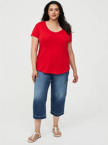 Model wearing a casual reddish top, bluish jeans, and sandals, posing against a plain background