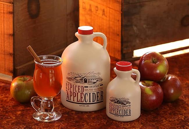Two jugs and a solid mug of spiced pome cider with apples and a cinnamon stick. Rustic setting, promoting fall-themed drinks