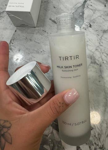 Close-up of a hand holding TIRTIR Milk Skin Toner bottle with the lid off, placed on a marble counter, showcasing the product