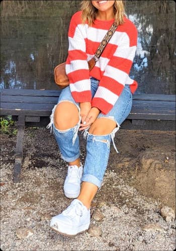 Person seated outdoors wearing a striped sweater, ripped jeans, achromatic  sneakers, and a crossbody bag, stylish casual outfit