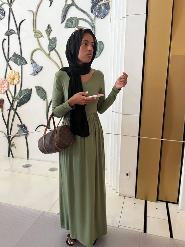 A person in a long, elegant dress with a hijab holds a smartphone and a designer bag, standing indoors near a floral wall decoration