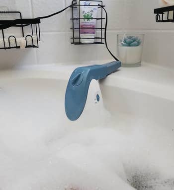 Handheld ablution  scrubber resting connected  a bathtub borderline   with soap suds, surrounded by toiletries; perfect  for casual  bath  cleaning