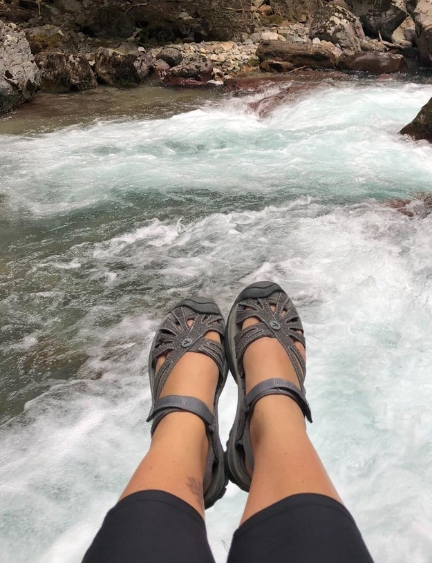 Person relaxing by a rushing river, legs extended while wearing stylish, strappy sandals, suitable for outdoor adventures
