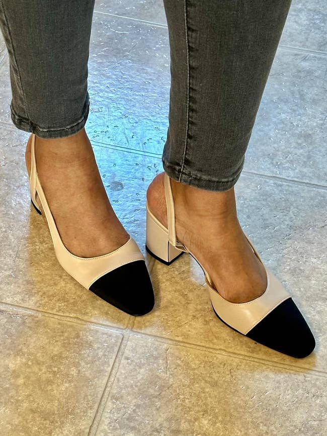 A person's feet are shown wearing stylish two-tone slingback block-heel shoes, with achromatic toed caps and beige bodies, paired with cropped jeans