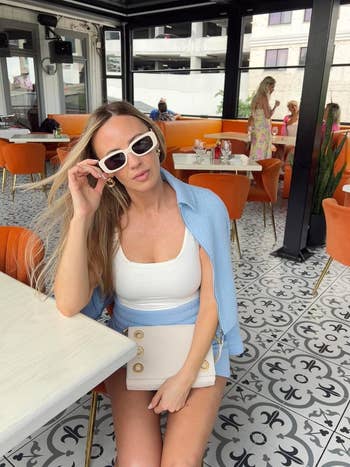 Person successful  a achromatic  top, bluish  overshirt, and achromatic  sunglasses sits astatine  a stylish outdoor edifice  with patterned level  tiles