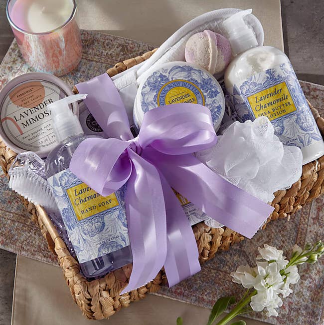 Brown wicker basket with large light purple bow filled with bottle of lavender scented hand soap, shea butter lotion, bath bomb, candle, foot brush, body butter, and white slippers