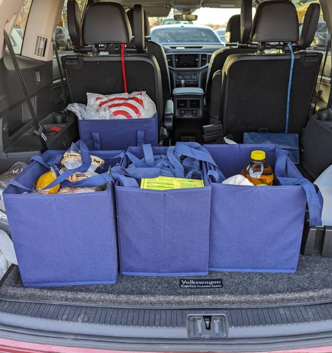 An open car trunk with multiple blue square-shaped shopping bags filled with assorted groceries