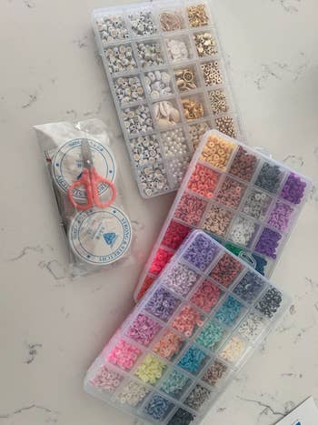 Assorted beads and jewelry-making supplies arranged on a table, including storage boxes with sections and a packet of small tools