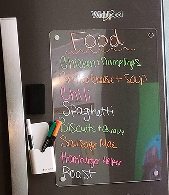 A clear rectangular dry erase board hung vertically on a fridge with an ingredients list