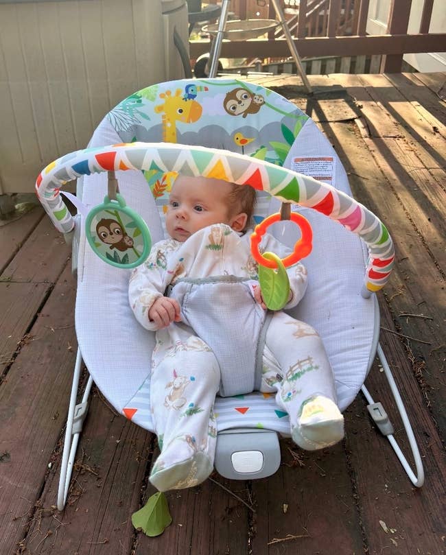 Baby successful  a onesie sits successful  an animal-themed bouncer chair, holding a colorful ring, outdoors connected  a woody  deck