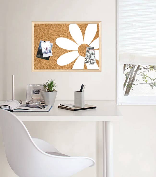 Modern white desk with chair, corkboard featuring a daisy design, and various office supplies by a window with sheer blinds