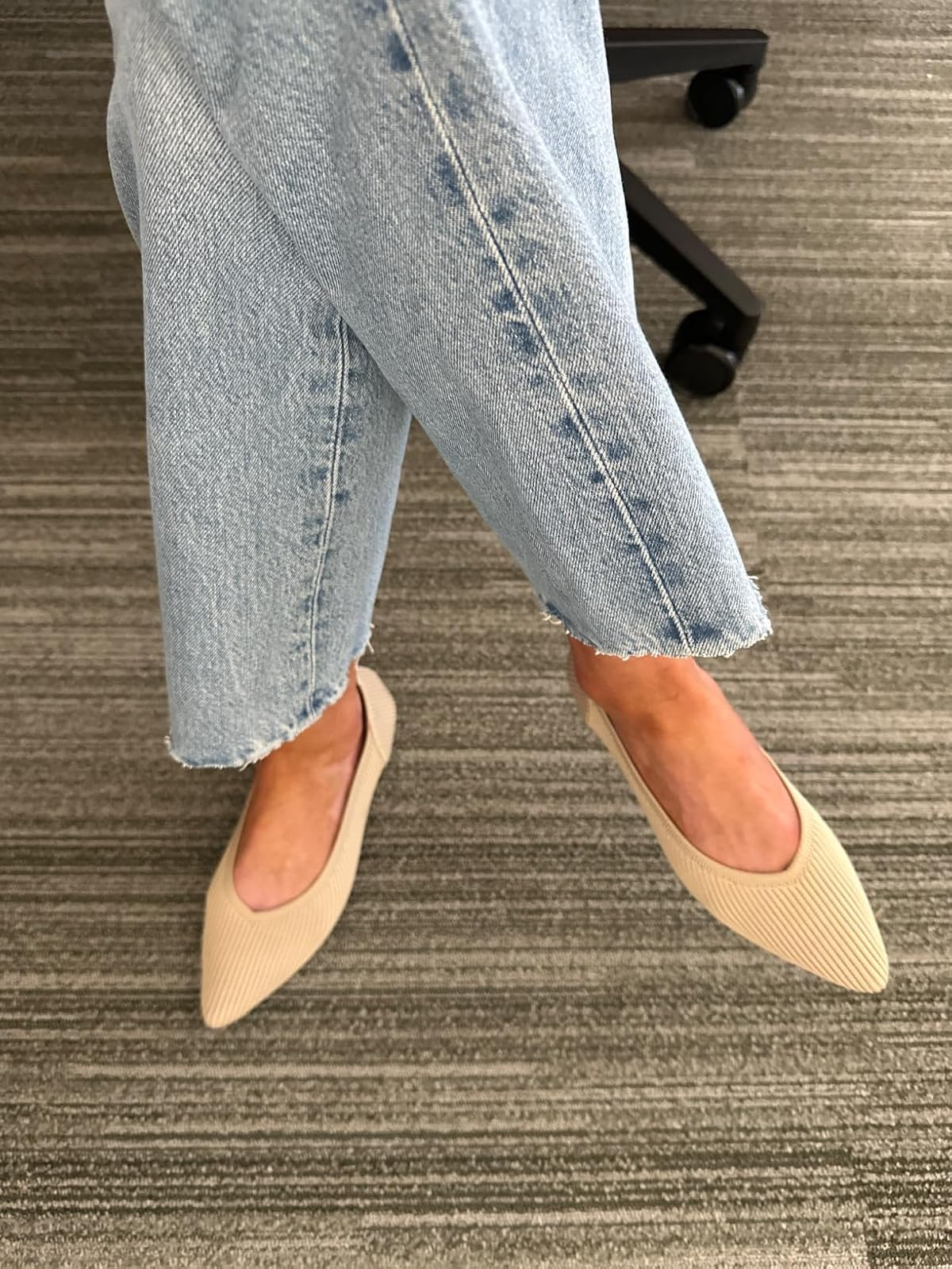 Person wearing light denim jeans with raw hems and beige pointed flats on a textured floor