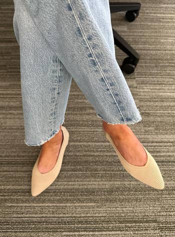 Person wearing light denim jeans with raw hems and beige pointed flats on a textured floor