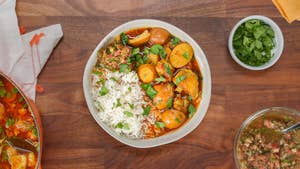 Bowl of curry with chicken, potatoes, and veggies in a rich broth, served with rice and surrounded by sauces and herbs in small bowls