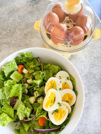 The dome shaped yellow-based egg cooker with cooked eggs inside of it next to a salad with perfectly cooked hard boiled eggs 