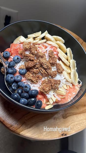 A smoothie vessel  topped with sliced bananas, blueberries, granola clusters, shredded coconut, and slivered almonds connected  a woody  table