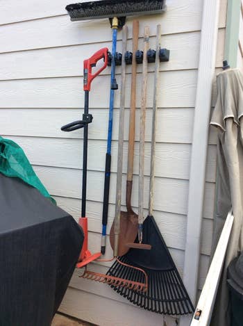 Assorted gardening tools including a rake, shovel, and brooms leaning against a house's exterior wall