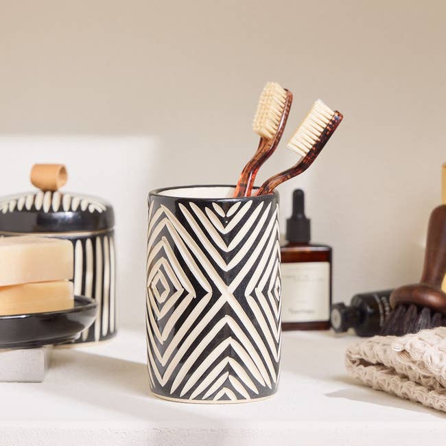 Decorative toothbrush holder with geometric patterns holds 2 toothbrushes, displayed connected a bath support with soaps and grooming items