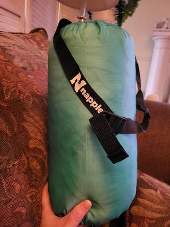 Person holding a teal pillow stuff sack with pillow inside and 'Napple' logo on the strap