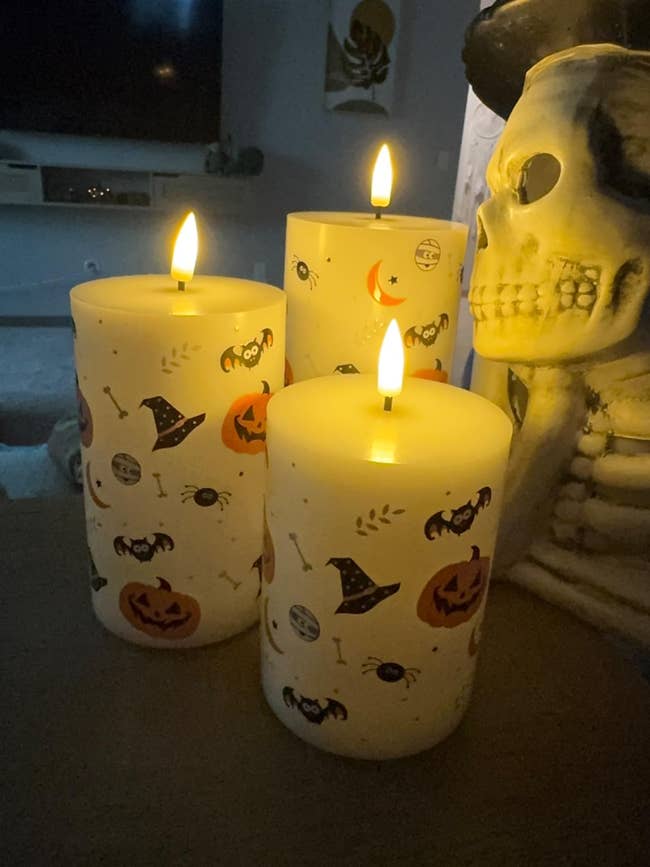 Three candles with Halloween-themed designs, including bats, pumpkins, and witch hats, placed adjacent  to a decorative skeleton figure