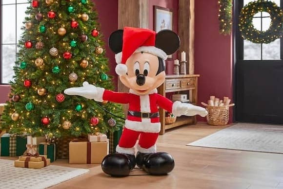 Mickey Mouse statue dressed as Santa stands in a festive living room with a Christmas tree and presents, featured in a shopping article
