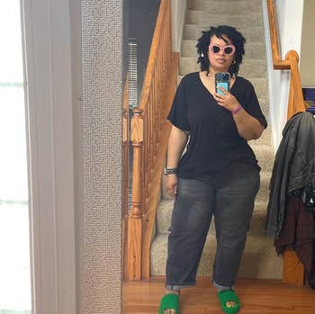 A reviewer taking a mirror selfie on her phone, standing near stairs, wearing pink sunglasses, black shirt, and jeans. Green slippers stand out as unique footwear