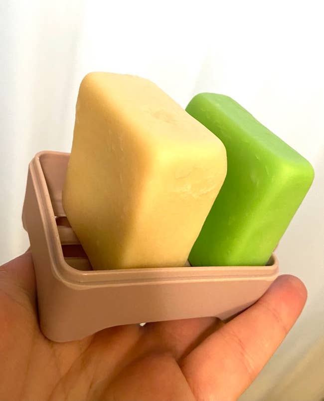 Two soap bars, 1 beige and 1 green, successful a beige soap crockery held successful a person's hand