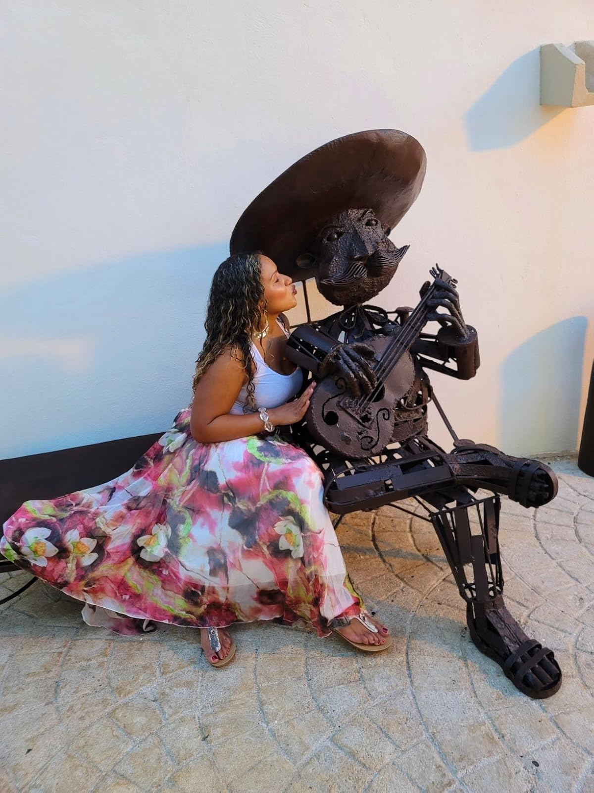 A reviewer in floral skirt poses with metal sculpture of a musician holding a guitar, playfully interacting outdoors