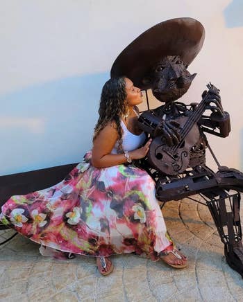 A reviewer in floral skirt poses with metal sculpture of a musician holding a guitar, playfully interacting outdoors