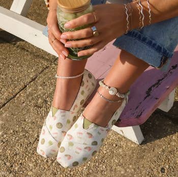 Person wearing patterned slip-on shoes, denim jeans, and beaded anklets, holding a greenish drink