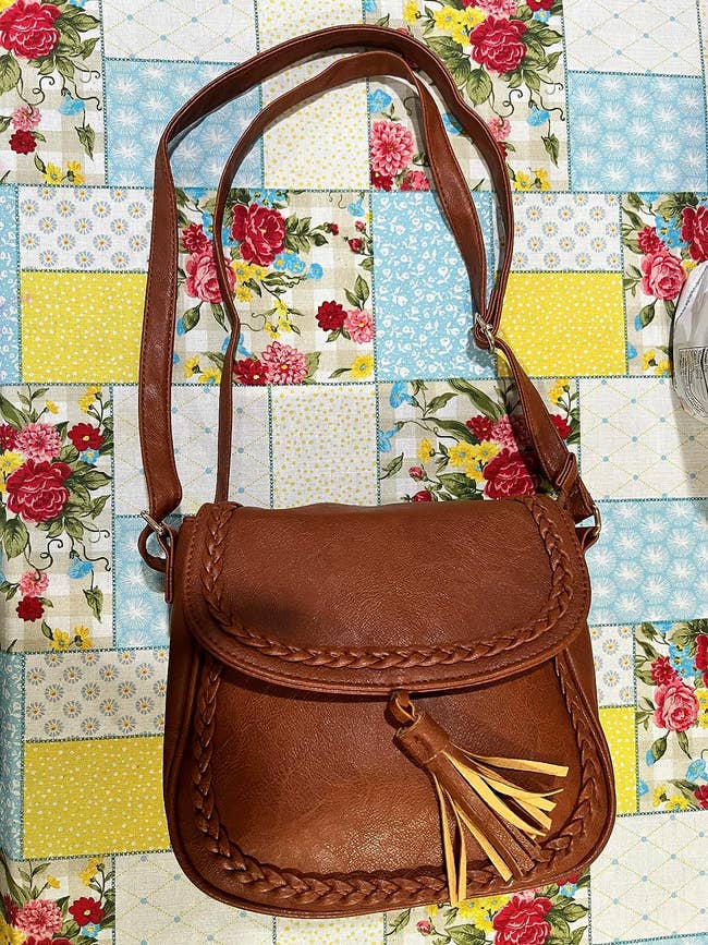 Brown leather crossbody bag with braided details and tassel accent rests on a floral and geometric patterned surface